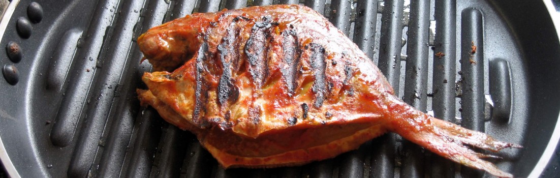 Whole grilled fish with charred grill marks and reddish glaze, resting on a black ridged grill pan